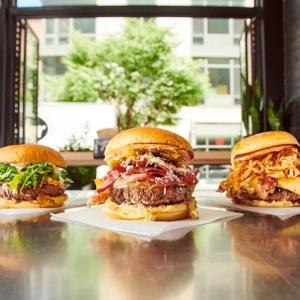 Three gourmet burgers from Black Tap Dallas displayed on a wooden table near a large window, topped with greens, crispy bacon, melted cheese, and fried onions.