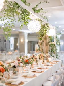 Vertical image of a sophisticated wedding reception with long, white-draped tables, floral centerpieces, candlelit place settings, and hanging greenery inside a refined event space.