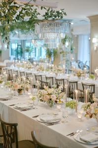Vertical image of a refined private event setup with long banquet tables, soft floral arrangements, sparkling chandeliers, glowing candlelight, and overhead greenery in a chic indoor venue.