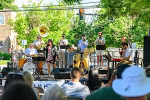Eight musicians perform on a stage under a shady tree canopy on Broad Street in Kennett Square. A woman in a floral dress sings with a crowd sitting attentively.