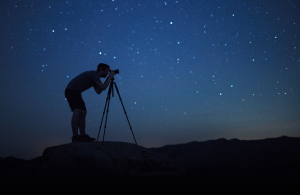 A photographer takes a picture with a camera and tripod silhouetted by a starry night sky. (Photo credit: Jack Fusco)