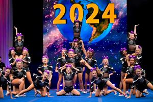 A Cheer Athletics team performs a vibrant pyramid formation during a 2024 competition, with athletes in black and silver uniforms accented by purple bows. The image captures the energy and precision that define Cheer Athletics programs nationwide, as the