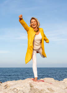 Clarissa Hoffmann standing in a powerful superhero pose on a rocky ocean cliff, embodying strength, confidence, and freedom against a backdrop of crashing waves and open sky.