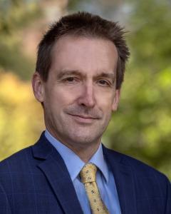 Dr Christopher Kiver. A man with short bown hair smiling at the camera. He wears a blue suit with a light blue shirt and a yellow tie.