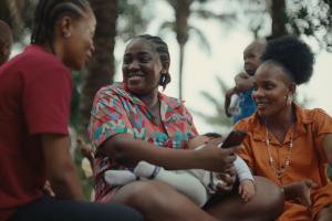 A smiling woman breastfeeds her baby while sitting with two other women in a park. They are chatting and laughing, surrounded by palm trees. One woman holds a phone and another cradles a toddler in the background.