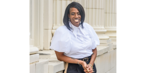 Anne Marie Otáñez smiling confidently in a white blouse and black pants, standing outside a classical building with white columns.