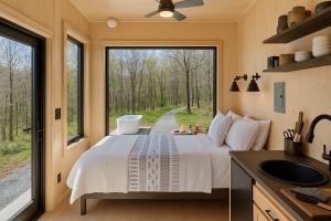 Minimalist cabin interior with a bed, modern kitchen, and expansive windows offering a forest view, featuring a cold plunge tub and fire pit outside.