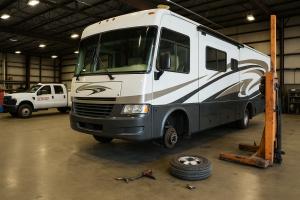 Large RV with front wheel removed inside a garage, alongside a service truck, tools, and hydraulic lift equipment.