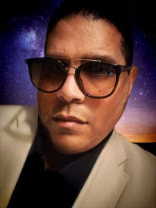 A picture of a smiling boydestiny wearing a tan blazer, black shirt and Gucci sunglasses.