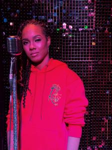 Ray Gifted Keys poses in a red hoodie with her crown-and-keys logo, holding a vintage mic on set of the “Wrong One” music video.