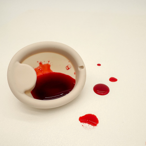 Top view of the Mimi™ menstrual disc partially filled with red fluid, showing its capacity and leakproof design, with blood-like drops on a white surface.