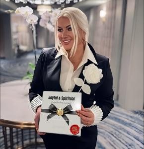 Julia Reno smiling and holding a copy of Joyful & Spiritual – Spring Edition, a beige hardcover book with a ribbon and gold seal, standing in an elegant hallway with soft lighting and white orchids in the background.