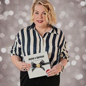 Dr. Sofia Kauko-Valli smiling while holding a copy of Joyful & Spiritual – Spring Edition, standing against a soft, sparkling background. The book cover features a cream design with a black ribbon and gold emblem, reflecting the theme of faith and renewal