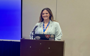 Female clinician in a navy blouse stands at a podium, gesturing toward a projected slide during a conference breakout session.
