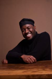 A handsome black man smiling with arms folded in dark clothing and hat.