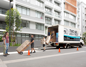 Image depicts a Two Men and a Truck van parked outside a unit block.  There are 3 removalists carrying moving boxes while a young couple watch.