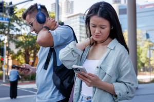 Image of young women standing outside, neck bent downwards at her mobile phone, standing next to a young man with headphone on staring at his phone.