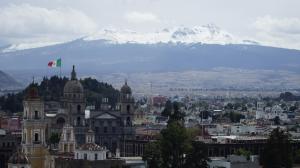 Panoramic view of Toluca, Mexico, featuring historic colonial architecture in the foreground, a large Mexican flag on a hilltop, and the snow-capped Nevado de Toluca volcano in the background under a cloudy sky.