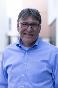 Kerry Brozyna, President of the Leather and Hide Council of America, smiling outdoors in a blue button-down shirt with glasses, short brown hair, and a relaxed demeanor. The background is softly blurred, showing light-toned buildings and natural lighting.