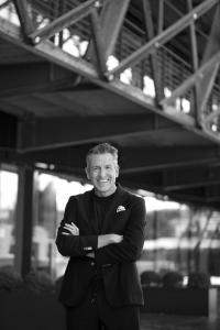Christopher Koerber, Managing Director of Hugo Boss Ticino and judge for the 2025 Real Leather. Stay Different. International Student Design Competition, smiling with arms crossed in a black suit and dark shirt. Photographed in black and white beneath a s