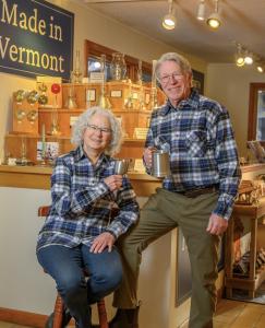 Older couple in plaid shirts smiling with pewter mugs in a Vermont shop showcasing handmade items