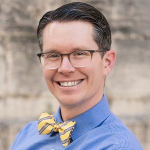 Shawn Hymel smiling, wearing glasses, a blue shirt, and a yellow striped bow tie, standing in front of a neutral background.
