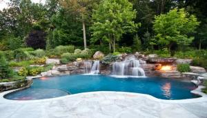 Luxurious backyard pool with cascading waterfall, surrounded by lush greenery and stone accents. A fire pit adds warmth and ambiance to the tranquil setting.