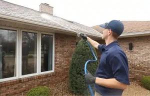 Man in blue cleaning brick house roof with a hose. He wears gloves and a cap, focused on task.