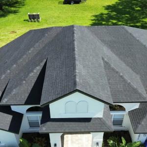Aerial view of a house with a dark, shingled roof and white walls, set against a lush green lawn.