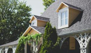 A charming house with a pitched roof, two dormer windows, and beige siding is surrounded by lush green trees.