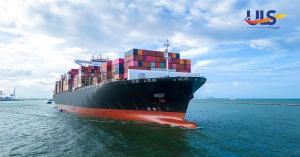 Large container ship carrying multicolored freight containers approaching port under a partly cloudy sky.
