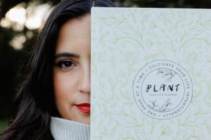 Stella Amador holding the PLANT Your Life Planner partially covering her face, with soft natural light and greenery in the background.