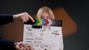 A clapperboard held in front of the camera on a film set, showing “Scene 5, Take 1” for a BTS shoot. The director is listed as Alex and the camera operator as John. A person with blonde hair is blurred in the background.