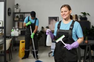 Female janitor in a black apron and blue uniform cleans office windows with a spray bottle and squeegee, while male coworker mops the floor behind her.
