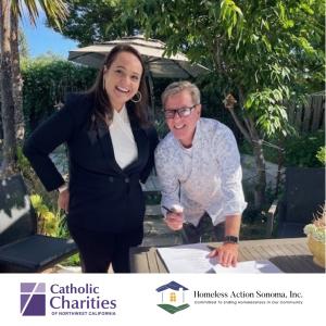 Jennielynn Holmes and James Whittaker stand at a table smiling and signing documents to formalize the partnership between Catholic Charities and Homeless Action Sonoma.