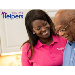 Smiling Senior Helpers caregiver in a bright pink polo shirt assisting an elderly man in a kitchen setting, with the Senior Helpers logo visible in the top left corner.