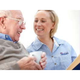 Senior Helpers caregiver in a light blue uniform smiling at an elderly man holding a white mug, creating a warm and friendly interaction in a bright indoor setting