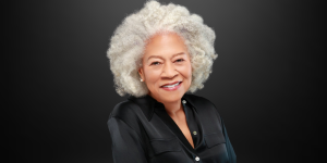 Dr. Andrea D. Sullivan smiling warmly against a dark gray background. She has white curly hair, wears a black button-down blouse, and exudes confidence and approachability.
