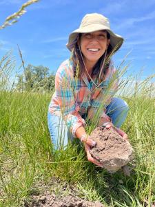 A smiling woman in a sunhat and plaid shirt kneels in tall grass, holding a clump of soil, as part of Boomitra’s regenerative grazing project in Argentina.