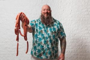 Chef Ralph Perrazzo smiling and holding a large handful of Snap-O-Razzo hot dogs in front of a white brick wall.