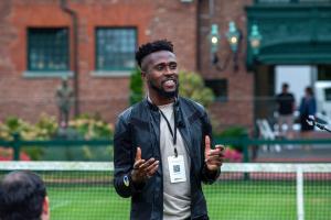 Tim Salau, Mr. FoW wearing a leather jacket and conference badge speaks to a group outdoors, using hand gestures and a microphone headset, with a tennis court and historic building in the background.