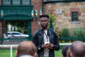 Tim Salau wearing a leather jacket and conference badge speaks to a group outdoors, using hand gestures and a microphone headset, with a tennis court and historic building in the background.