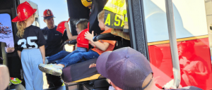 A child in a fire truck seat, wearing a fire helmet, engages with firefighters, while others observe nearby in a sunny outdoor setting.