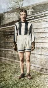 Colorized photo of Pat White standing in racing attire—striped jersey, shorts, gloves, and leather shoes—on a grassy field with wooden bleachers in the background.