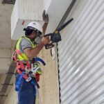 Technician working on scaffolding to repair or maintain an industrial sectional garage door with multiple black vent panels in a warehouse - Garage Door Guys Perth