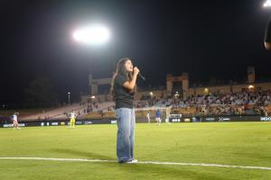 A young girl sings into a microphone on a soccer field at night under bright stadium lights, with spectators in the stands and players warming up in the background.