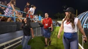 Teen artist Sofia Pineda, wearing a black LV Lights FC shirt and jeans, smiles and reaches up to high-five a young girl in the stands as other children cheer from behind the railing. Adults, including a woman in a white top and a man in a red shirt, walk