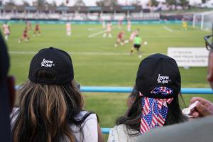 Two spectators wearing black “Learn The Arts!®” caps sit in the stands watching a live soccer match. One has long highlighted hair, and the other wears a patriotic red, white, and blue scarf under the cap. The game plays out on a green field in the backgr