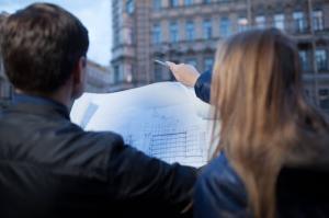 The male engineer holds the blueprints while his female colleague points at a historic building during documentation work