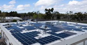 A commercial solar system stretches out across the flat roof of a business building. In the background there are trees, a small airport, and a sky with clouds.